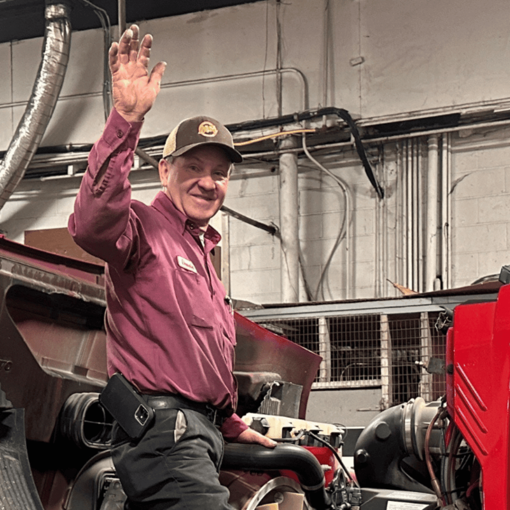 Long-time M&L Truck Service diesel mechanic and service manager Donald Benton waves hello, standing on the fender of a semi truck with the engine compartment open for repair.